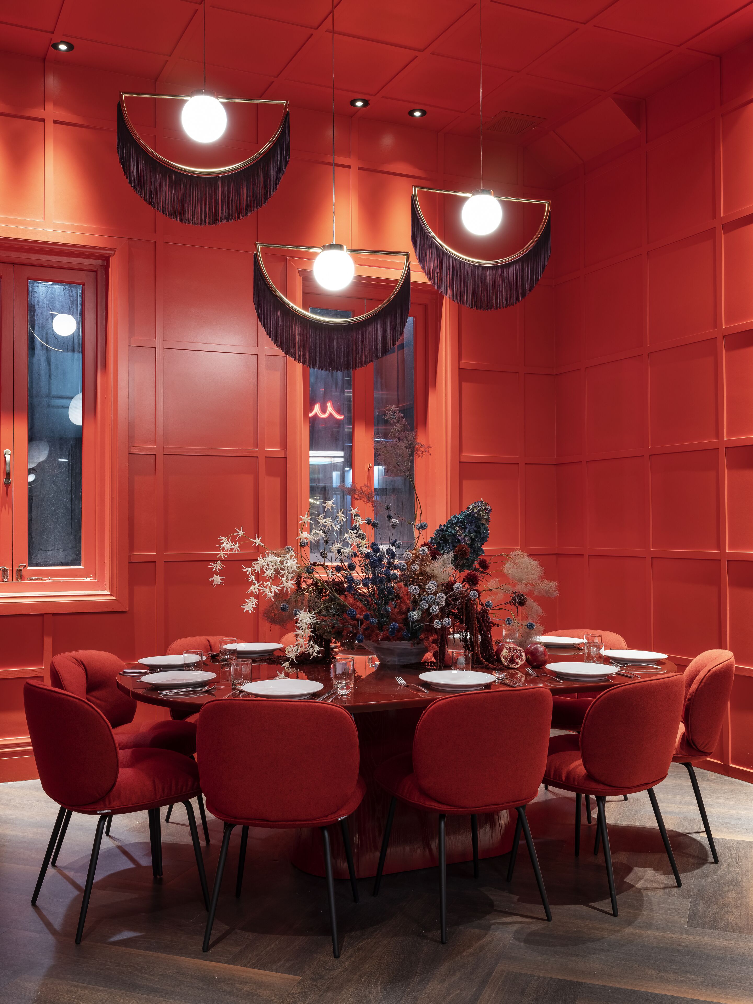 A dining area with a red color scheme, including walls, chairs, and table. There are three hanging pendant lights and a vase with flowers on the table.