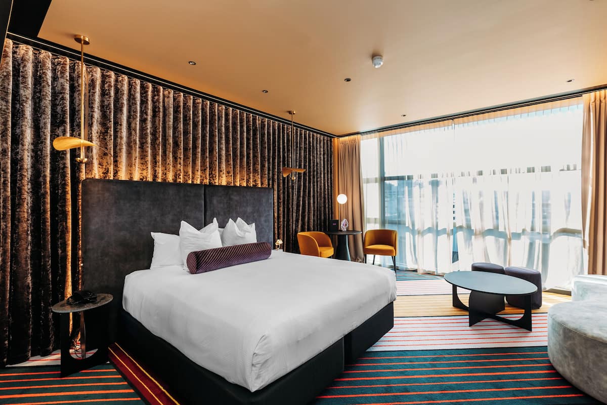 A modern hotel room with a large bed, two chairs, a small table, and a view of the city.