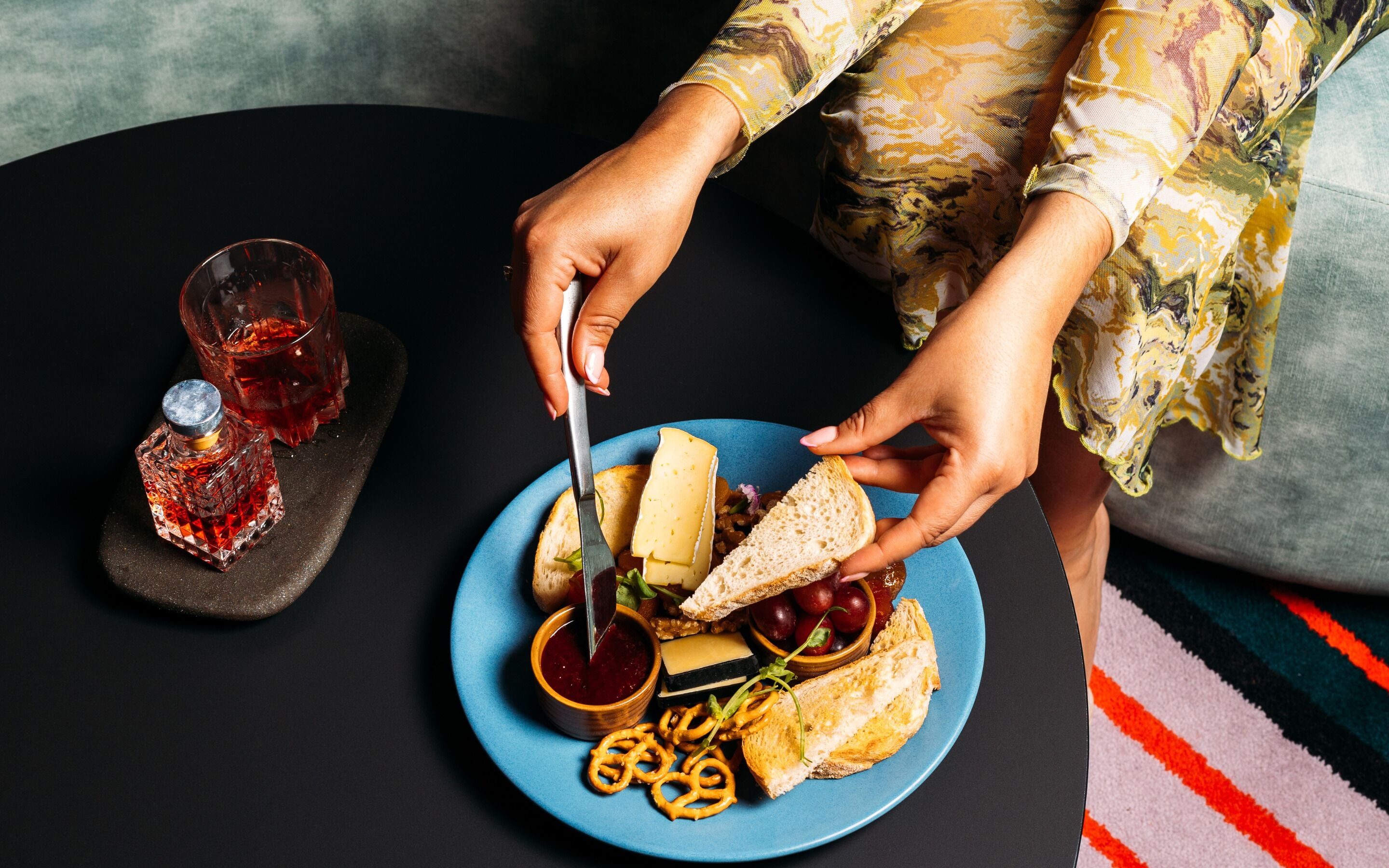 A person serving themselves a plate of cheese, bread, grapes, and pretzels from a blue plate on a black table.