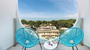 Ein Balkon mit zwei türkisfarbenen Stühlen, einem kleinen Tisch mit einem Buch und Sonnenbrille, und Blick auf eine Stadt mit Gebäuden und Bäumen.