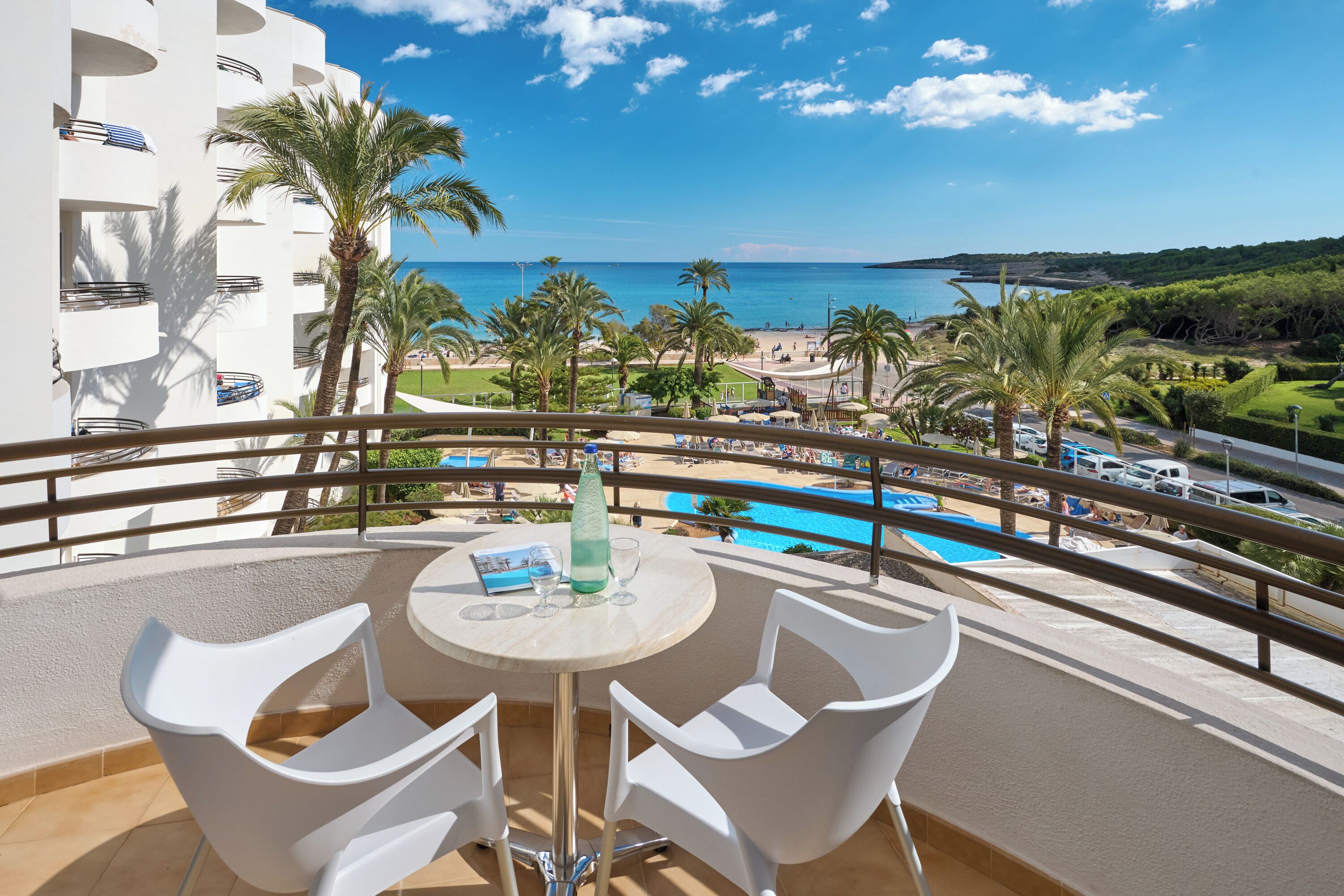Un balcón con sillas y una mesa blancos, con vista a una piscina y una playa rodeada de palmeras y un cielo azul despejado.