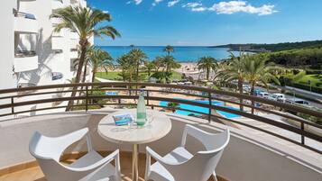 Un balcón con sillas y una mesa blancos, con vista a una piscina y una playa rodeada de palmeras y un cielo azul despejado.