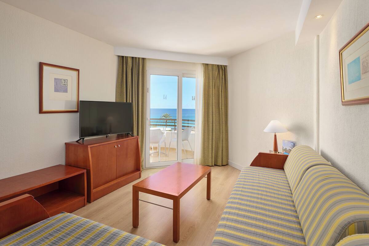 Hotelkamer met een bank, een houten tafel, een televisie en een balkon met uitzicht op zee.