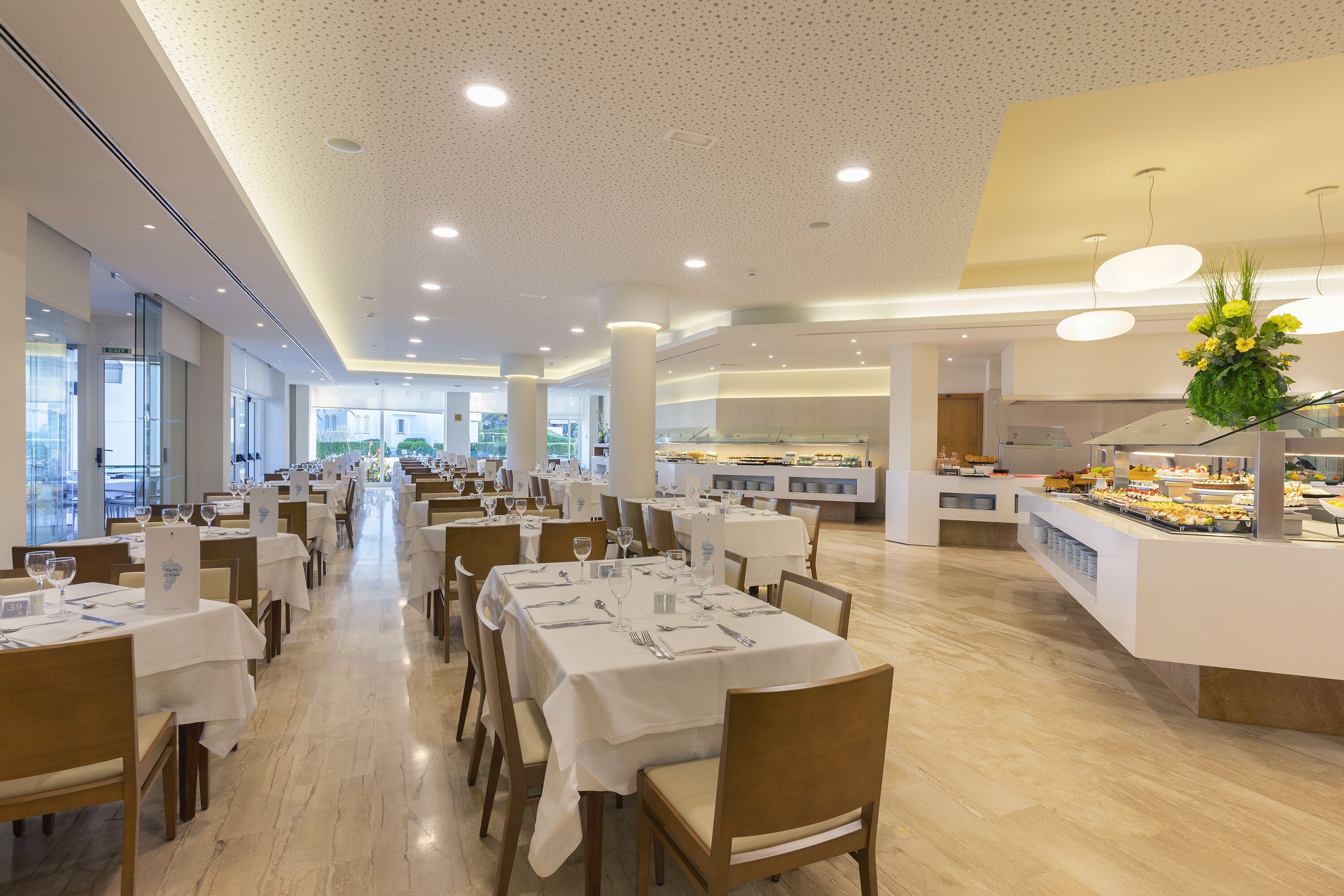 Een ruime eetruimte met meerdere tafels gedekt voor gasten, een centraal buffet en grote ramen die voor natuurlijk licht zorgen.