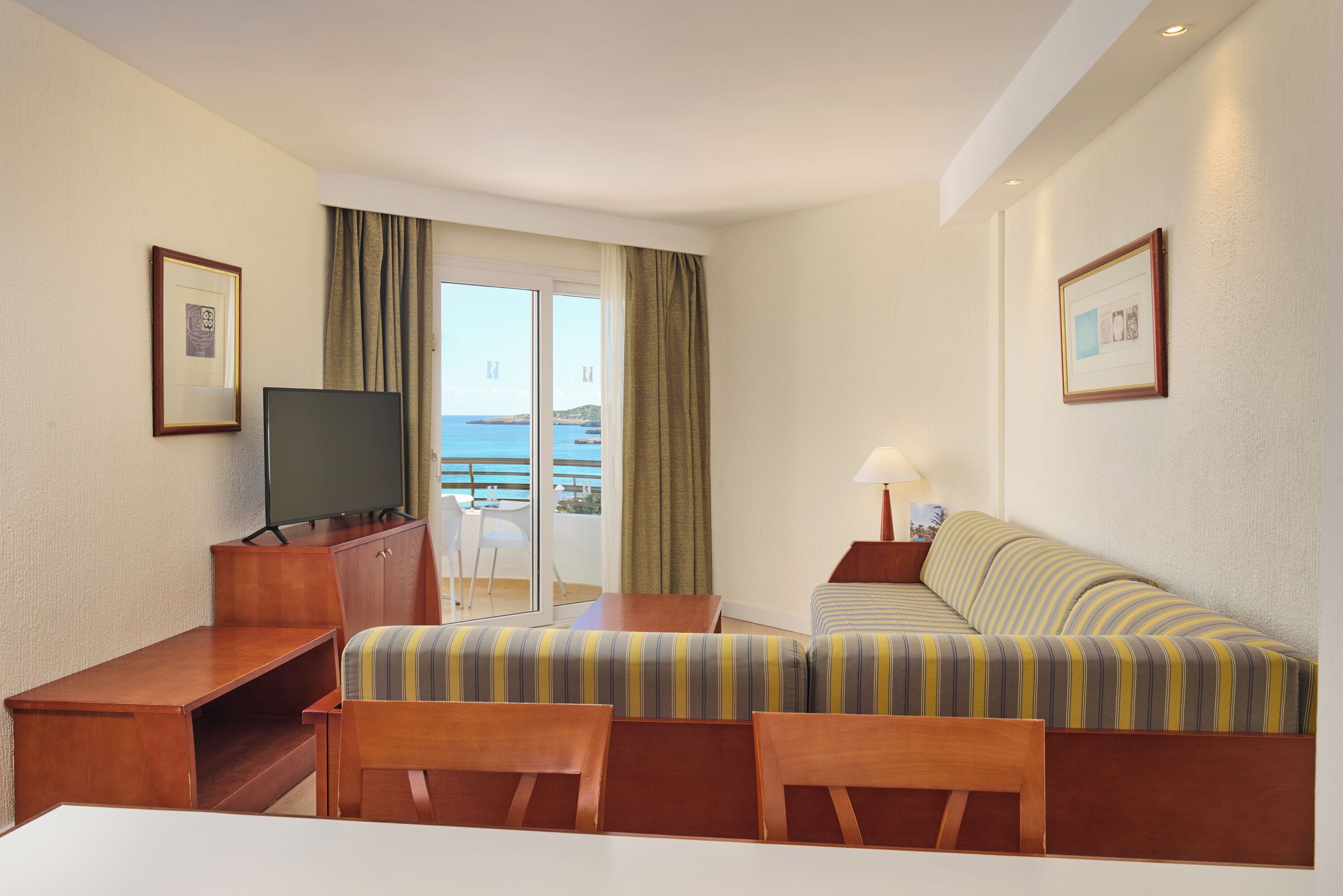 A hotel room with a bed, a TV, a desk, and a chair. There is a balcony with a view of the sea.