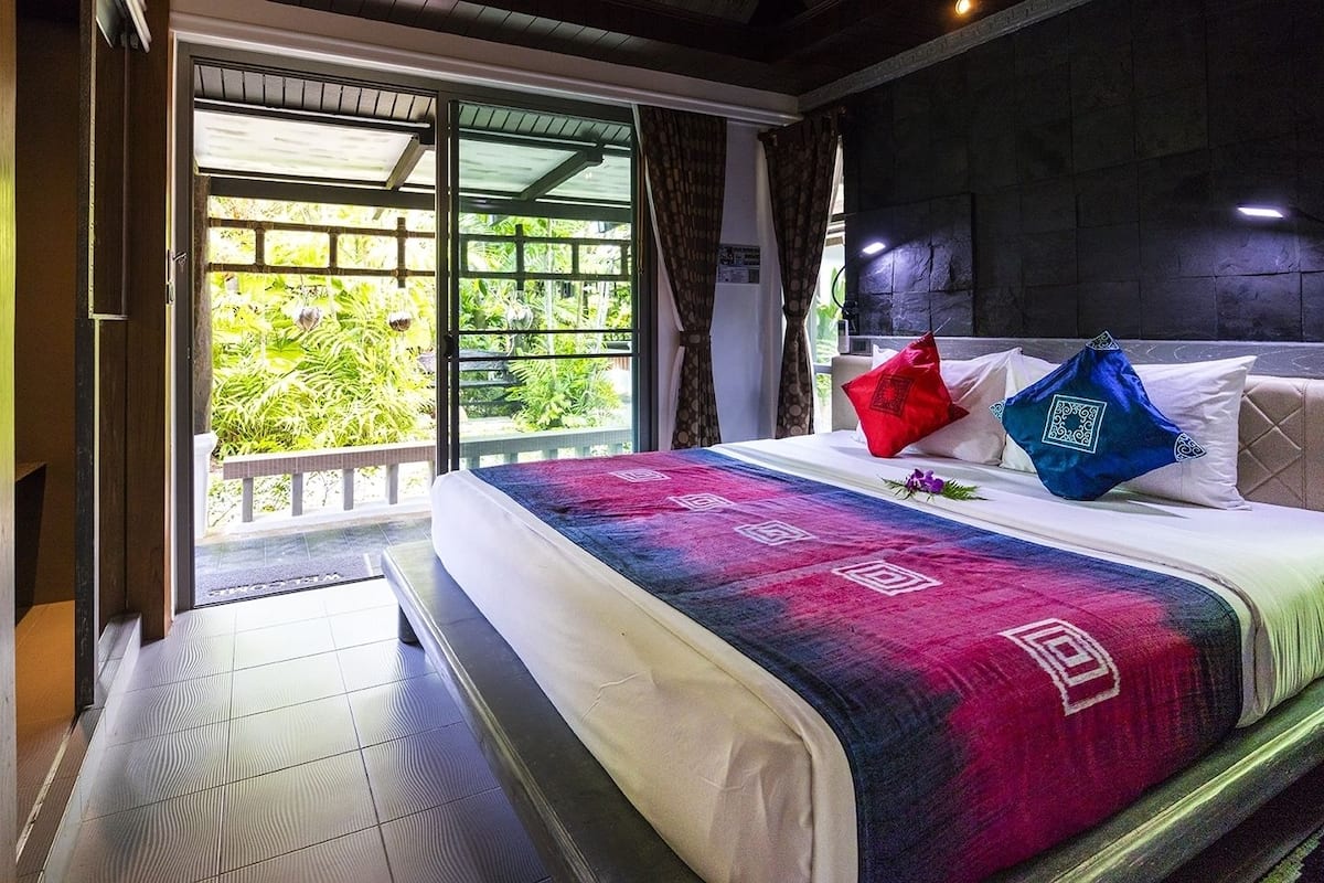 A hotel room with a bed, a view of greenery through the sliding glass door, and a dark stone wall.