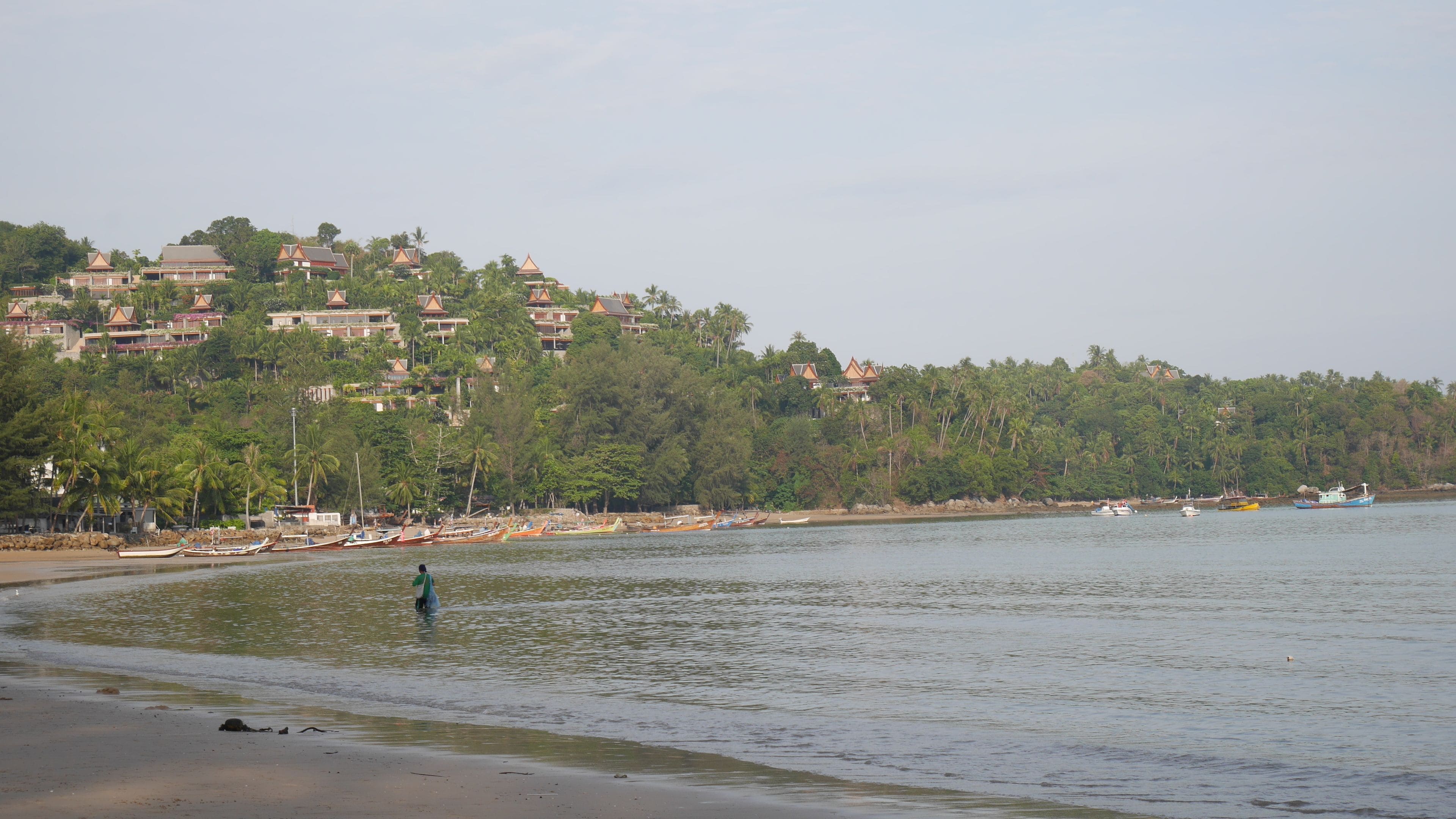 Una playa con barcos y una persona vadeando en el agua.