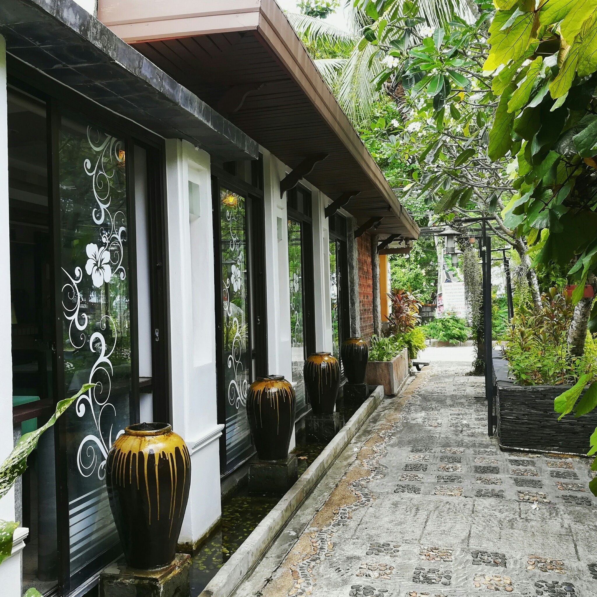 A corridor with a tiled floor, black and white patterned windows, and potted plants along the walls.