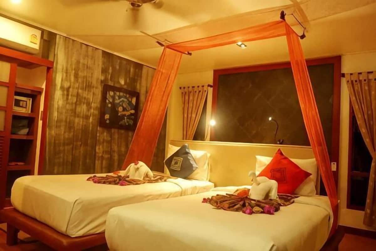 A room with two beds, a wooden headboard, a window with curtains, and a bookshelf.