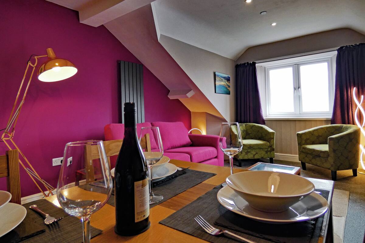 A dining area with a table set for two, a wine bottle, and glasses. There is a pink wall and a window with curtains.
