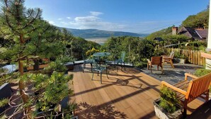 Una terraza de madera con mobiliario exterior, vista al mar y una cadena montañosa en la distancia.