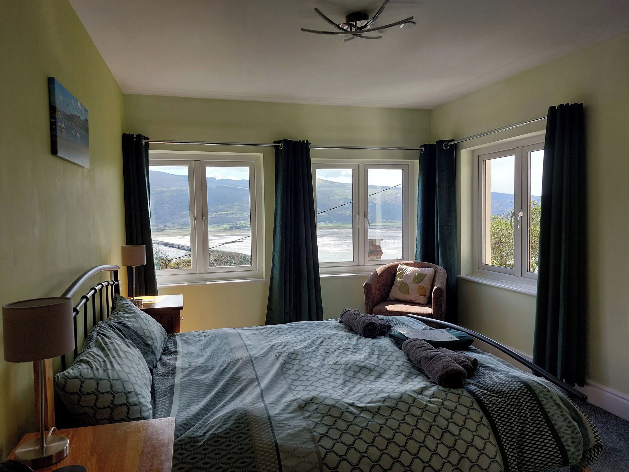 A bedroom with a bed, a chair, and two windows offering a view of a landscape.