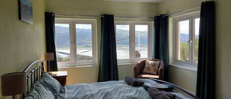 A bedroom with a bed, a chair, and two windows offering a view of a landscape.