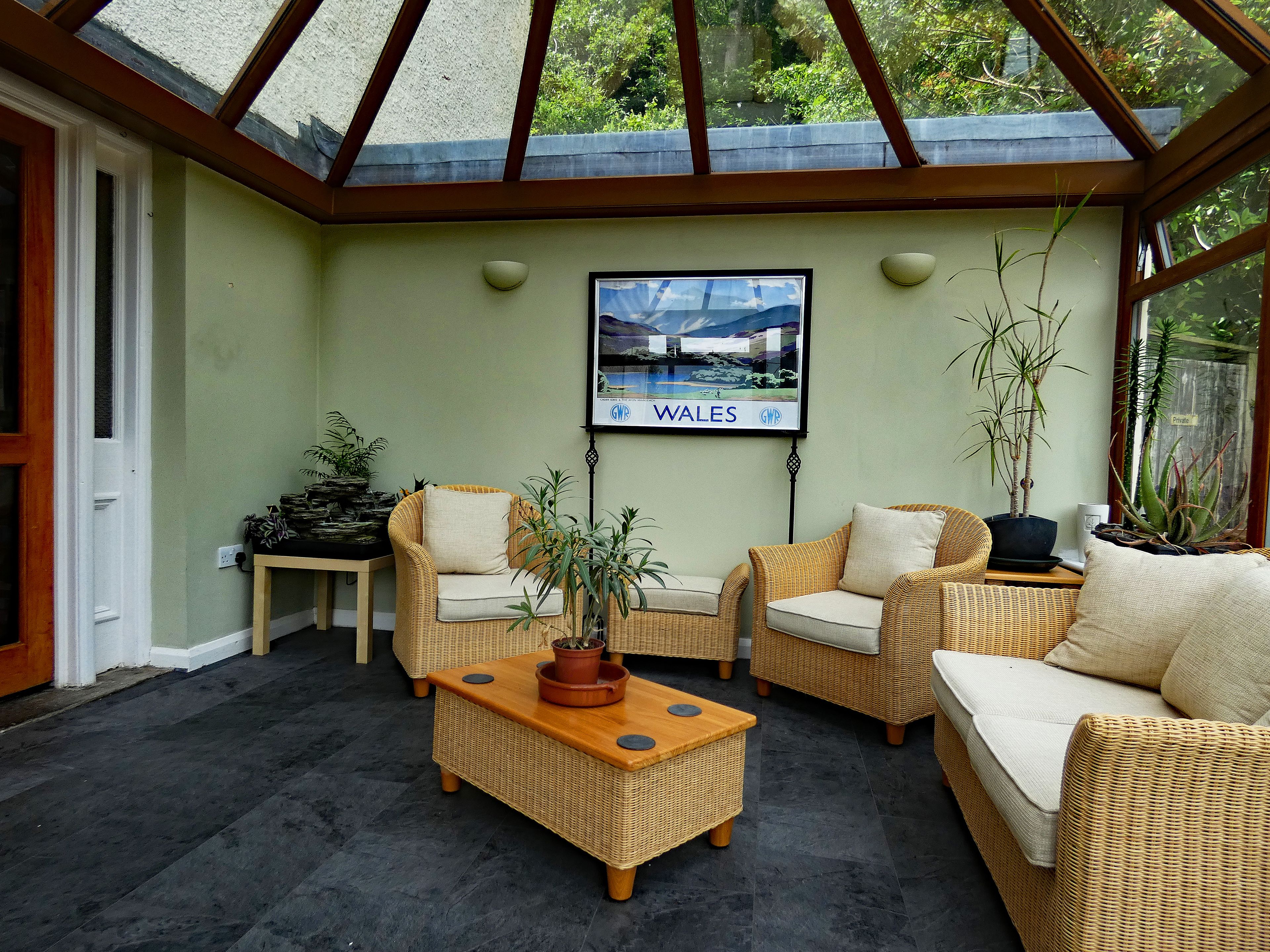 A sunlit patio with wicker furniture, a framed picture of Wales, and potted plants.