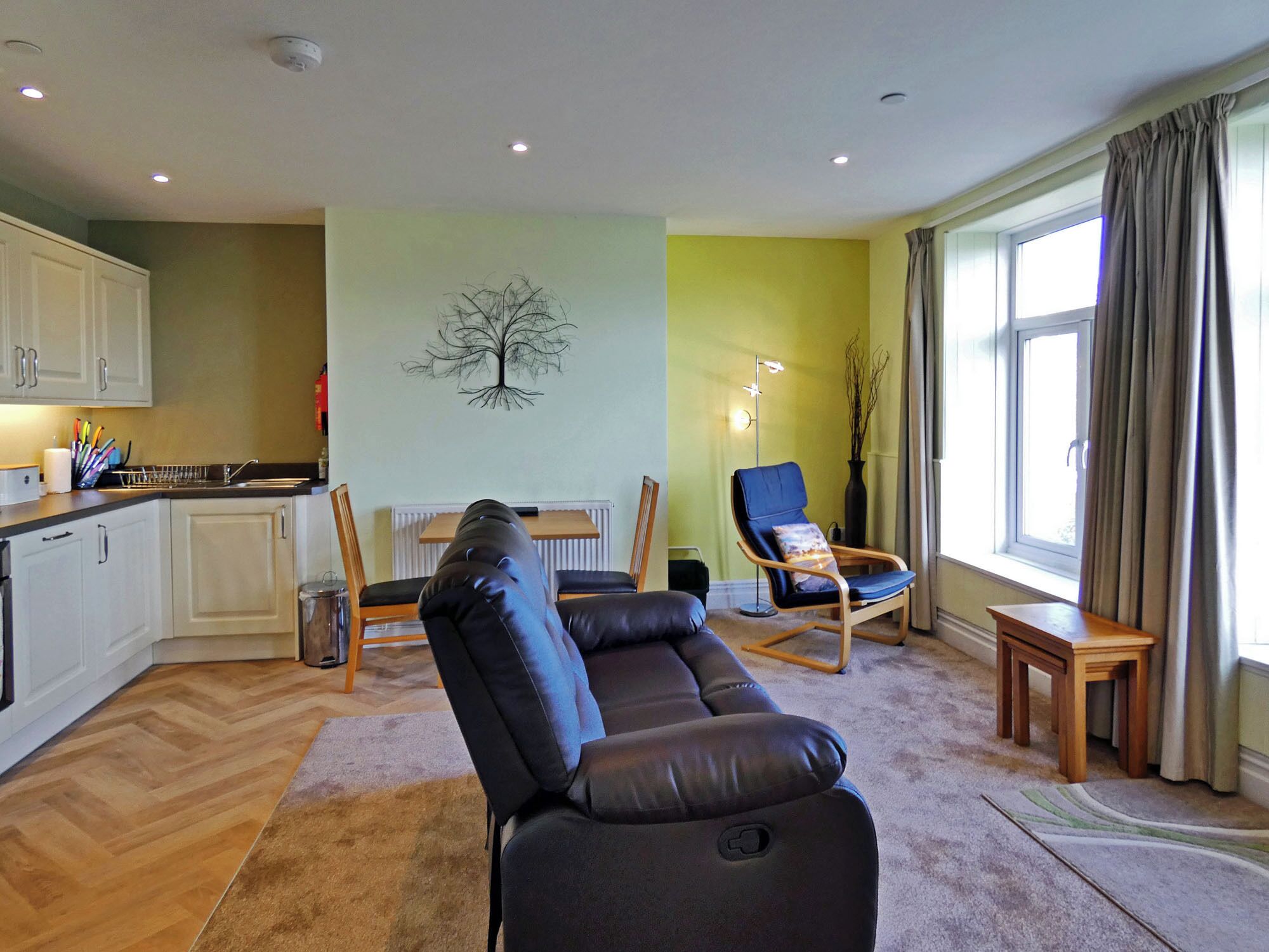 En moderne stue med en sort lædersofa, et spiseområde med bord og stole, et køkken med hvide skabe og et stort vindue med gardiner.