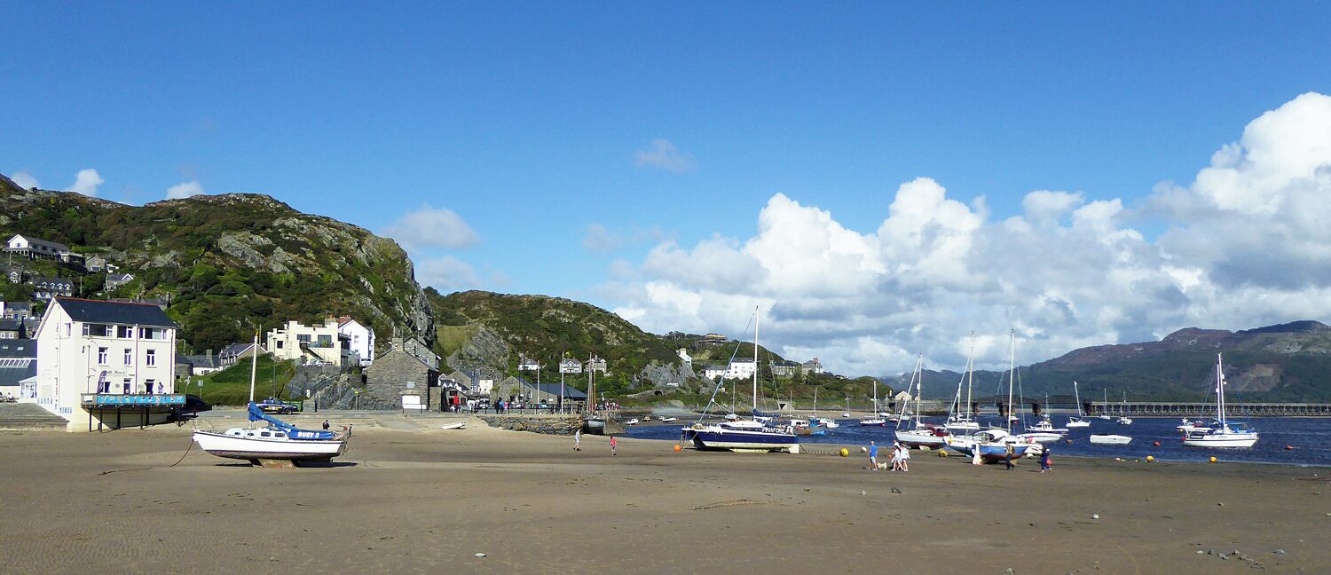 A coastal town with a sandy beach, boats, and buildings on a hillside.