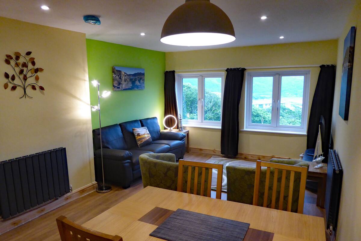 A living room with a sofa, a dining table, and a view of the outdoors through the windows.