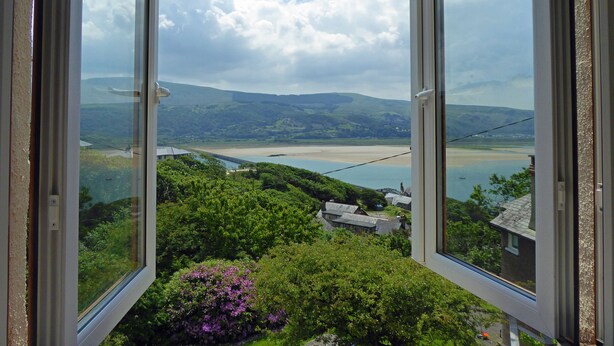 Vista desde una ventana sobre un paisaje costero con colinas, una playa y una extensión de agua.