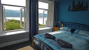 Un dormitorio con cama, mesita de noche, lámpara y vistas a un puente y vegetación a través de amplios ventanales.