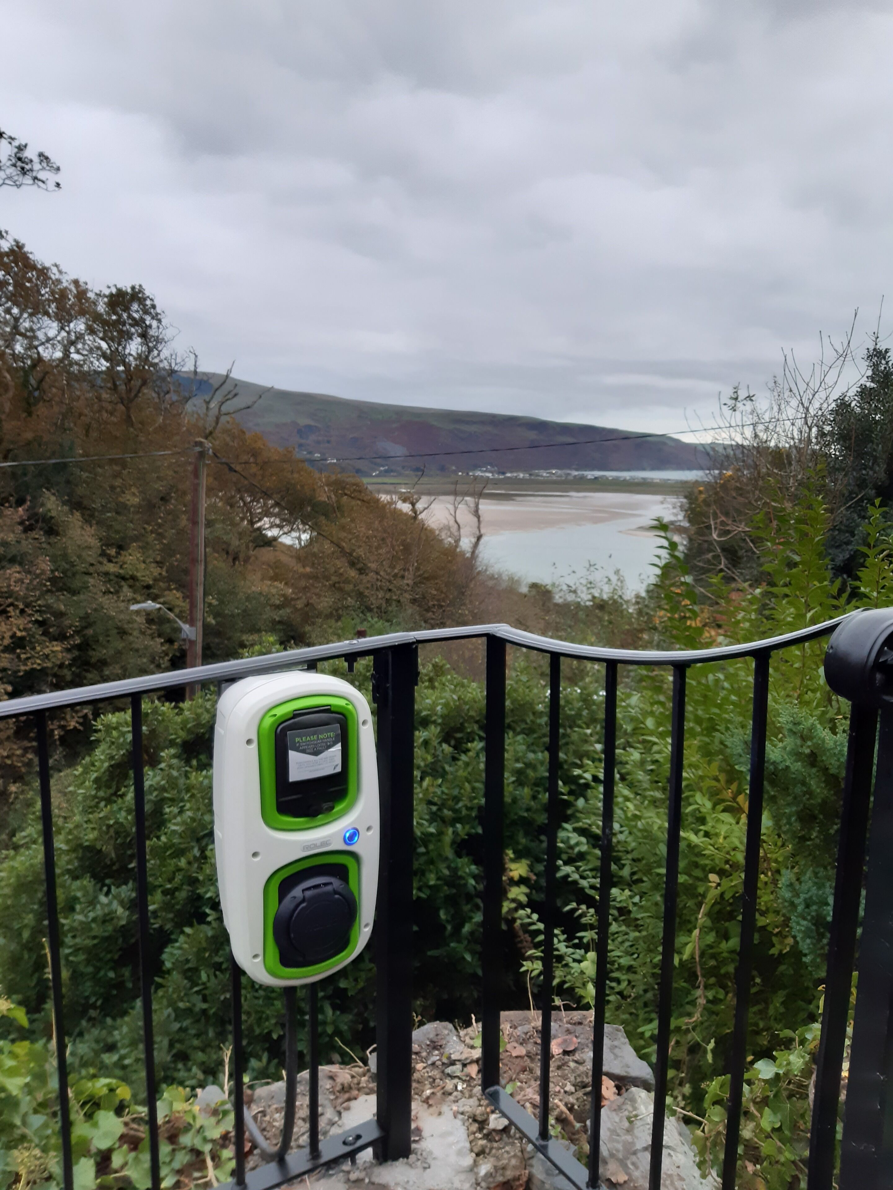A view of a coastal landscape with a green and white device attached to a black metal railing, overlooking a beach and hills.