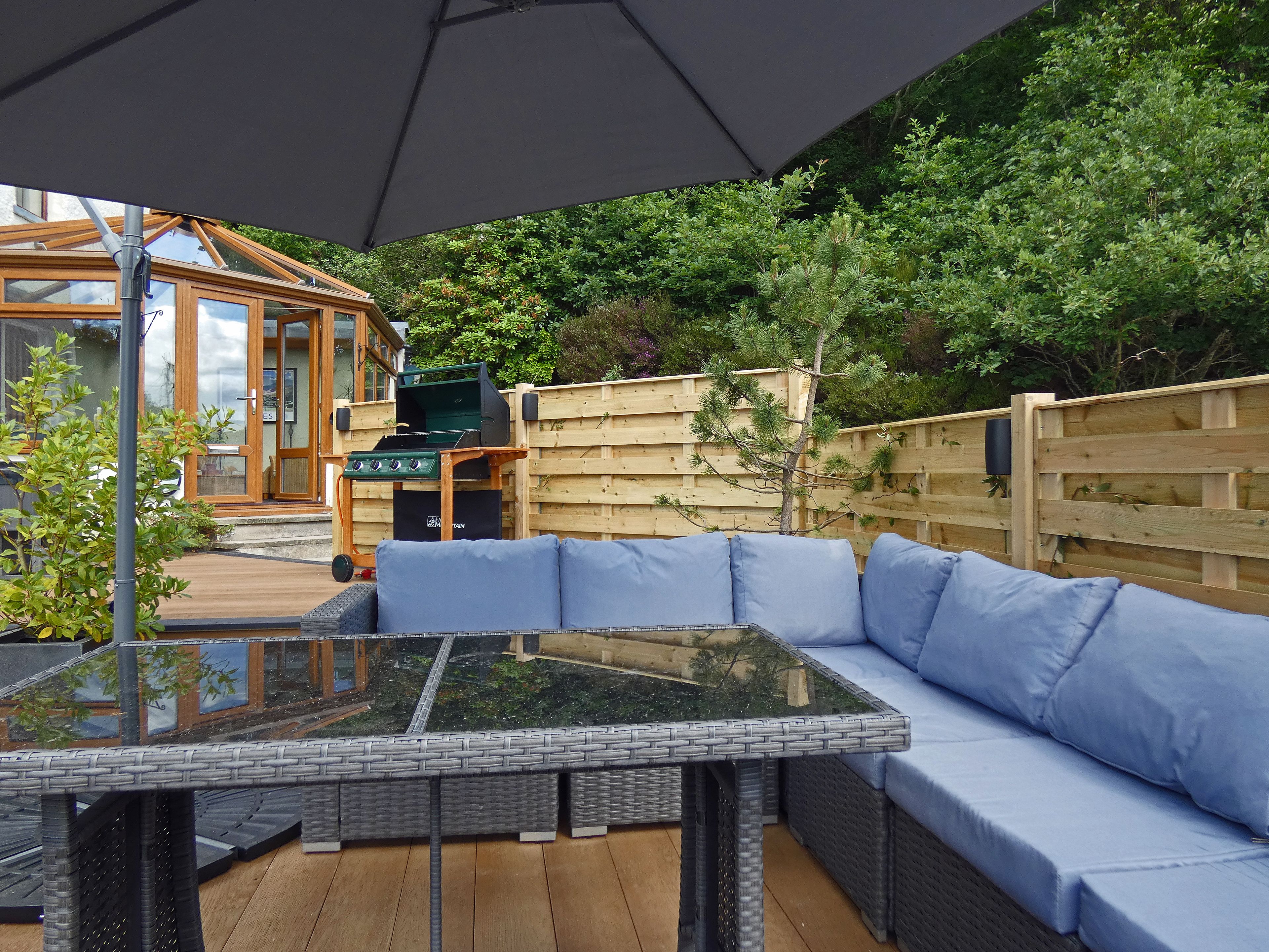 An outdoor seating area with a glass-top table and blue cushioned sofa, a barbecue grill, and a wooden deck.
