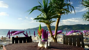 A beachside table with a view of the sea and mountains, surrounded by beach umbrellas and chairs. - Kudo Hotel (Patong)