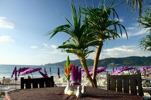 Een tafel aan het strand met uitzicht op zee en bergen, omringd door strandparasols en stoelen.