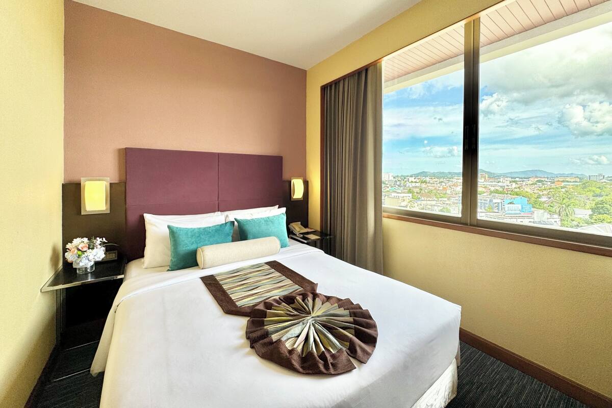 A hotel room with a large bed, a nightstand with a lamp, a window with a city view, and a headboard.