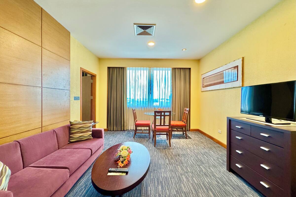 A hotel room with a sofa, a coffee table with a flower vase, a TV on a dresser, and a dining area with chairs and a table.