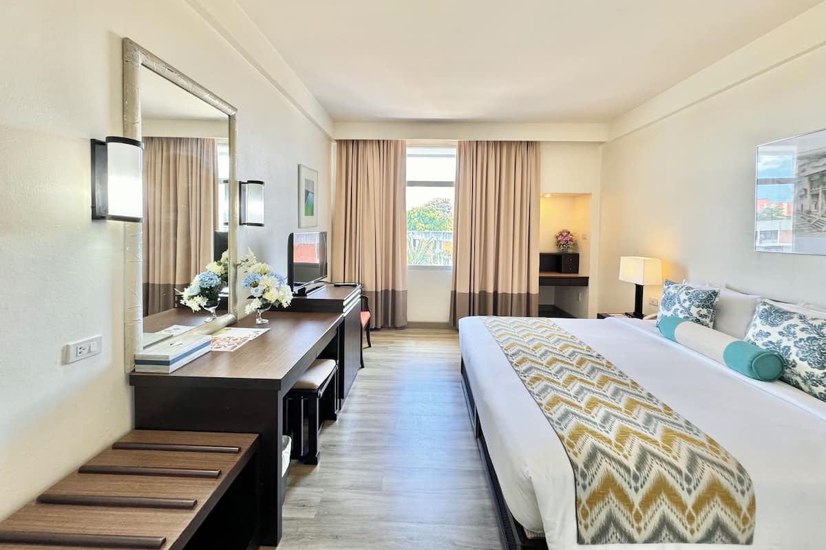 A hotel room with a bed, a desk with a chair, a TV, a lamp, and a window with curtains.