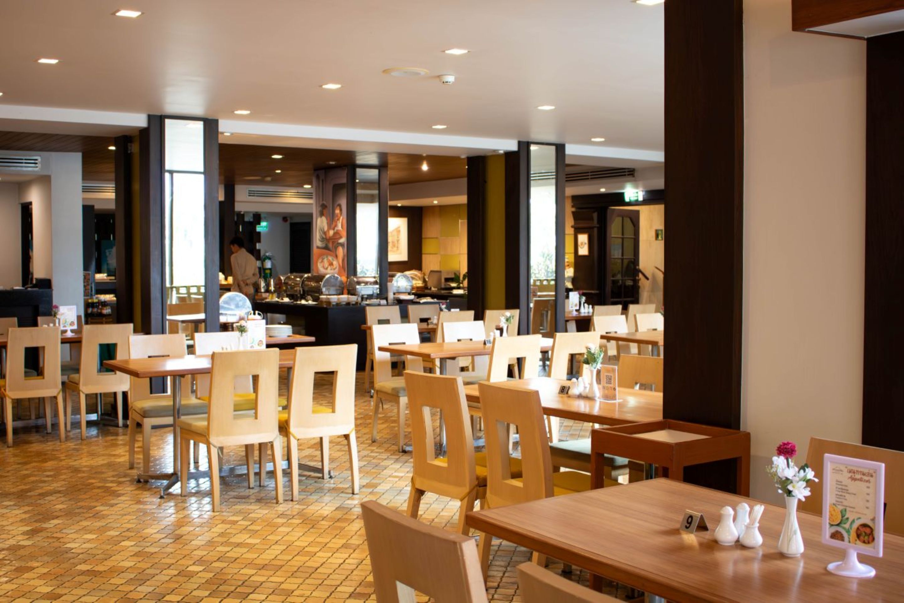Restaurante com mesas e cadeiras de madeira, chão de azulejos e uma pessoa ao fundo.