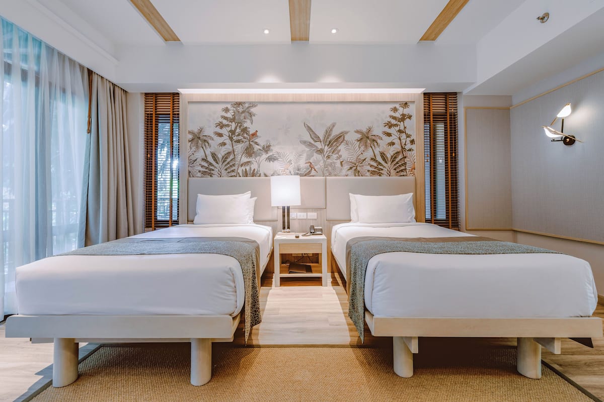 Une chambre d’hôtel avec deux lits, une table de chevet, une lampe fixée au mur et une grande fenêtre avec des rideaux.