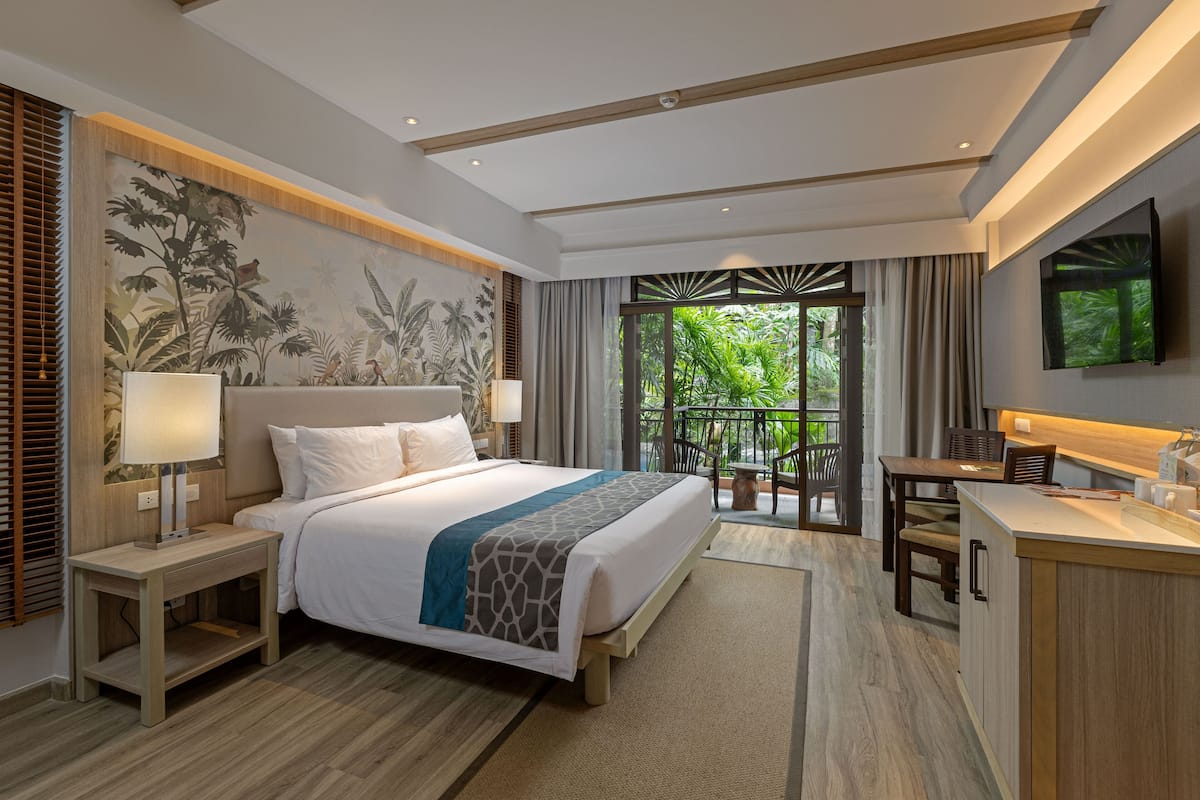 Une chambre d’hôtel moderne dotée d’un grand lit, de tables de chevet, d’un bureau et d’un balcon offrant une vue sur la verdure.