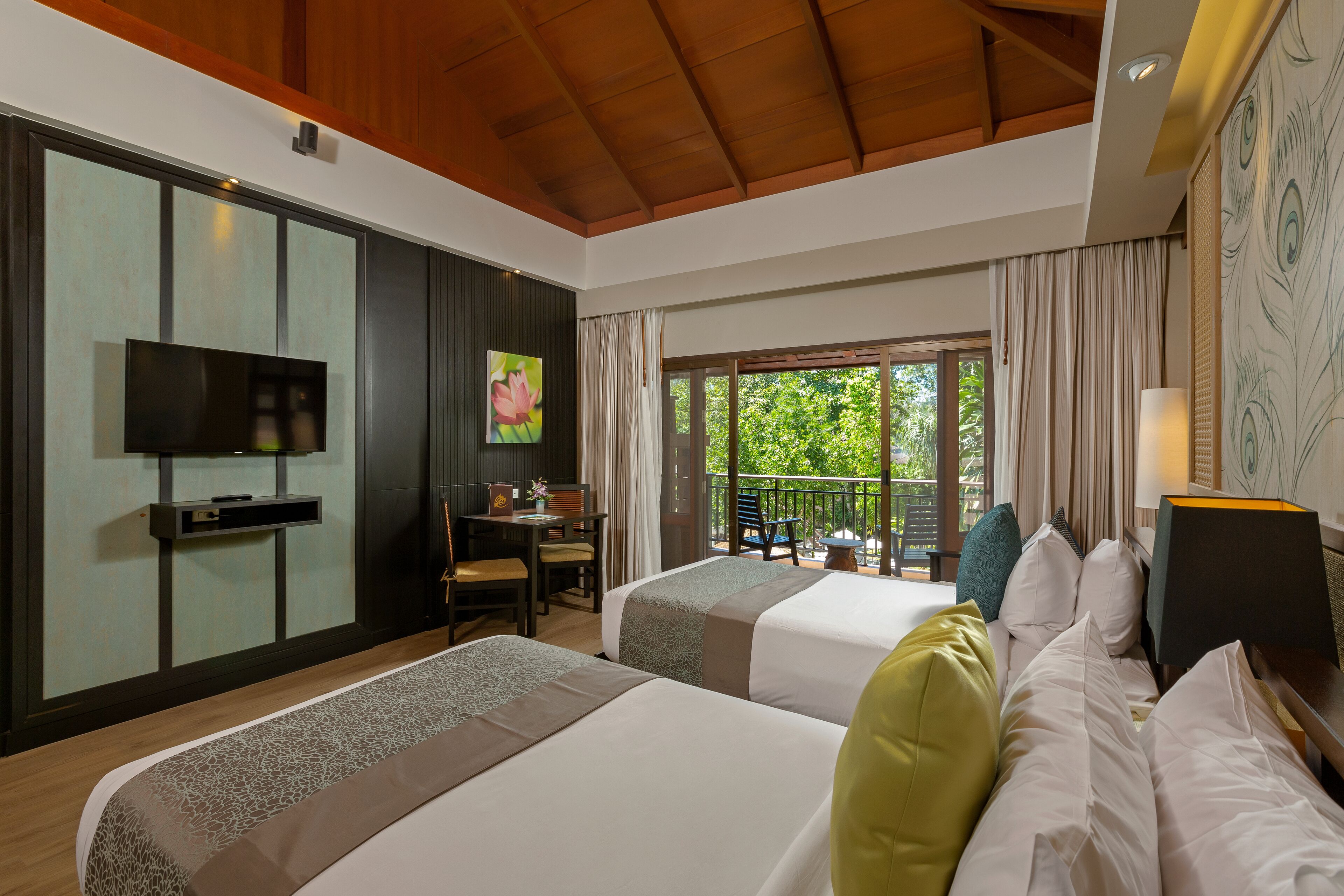 A hotel room with two beds, a desk, a TV, and a balcony with a view of greenery.