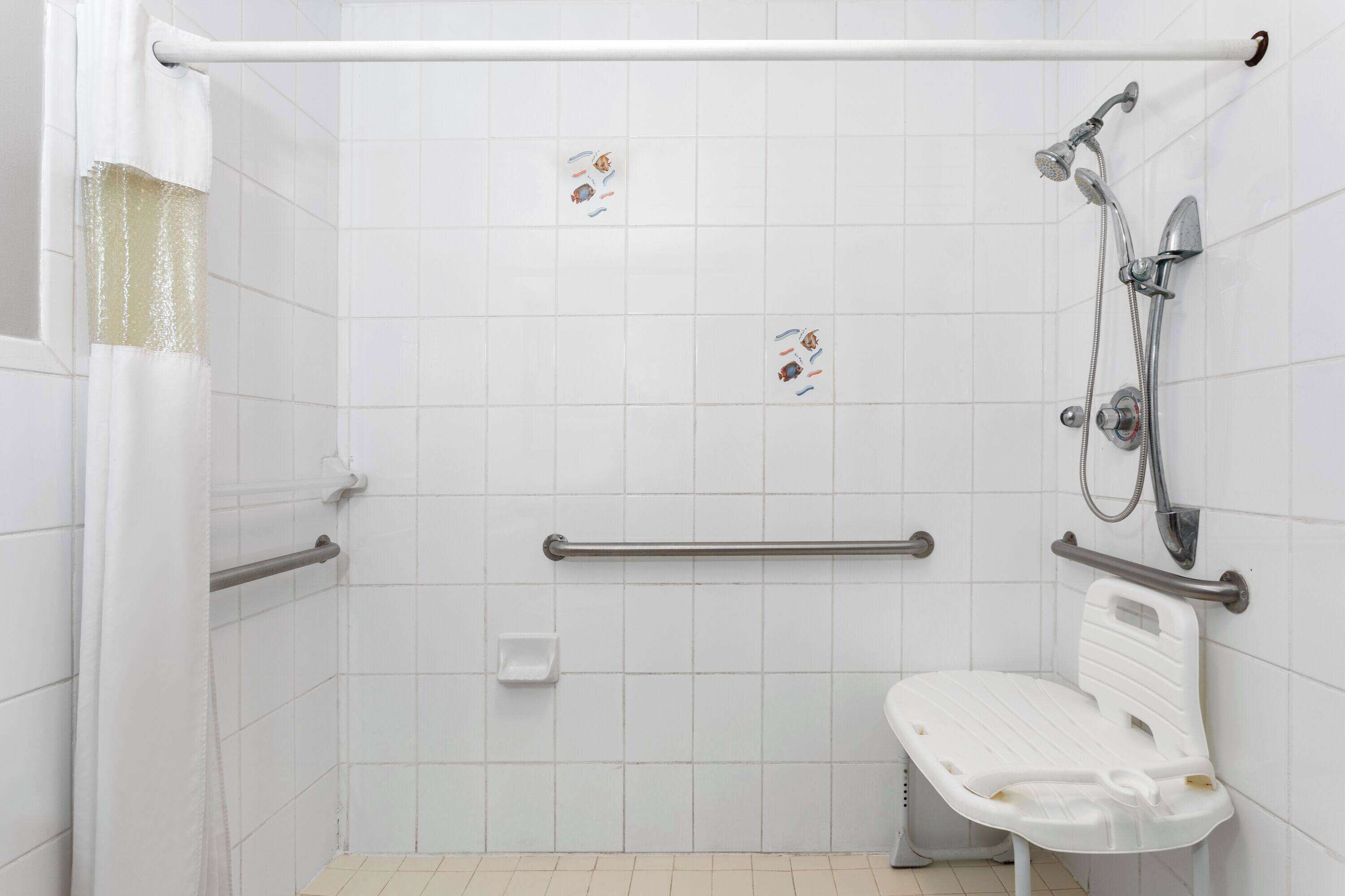 Une salle de bain avec des murs recouverts de carreaux blancs, une douche avec un pommeau de douche amovible, une barre d’appui et un banc blanc.