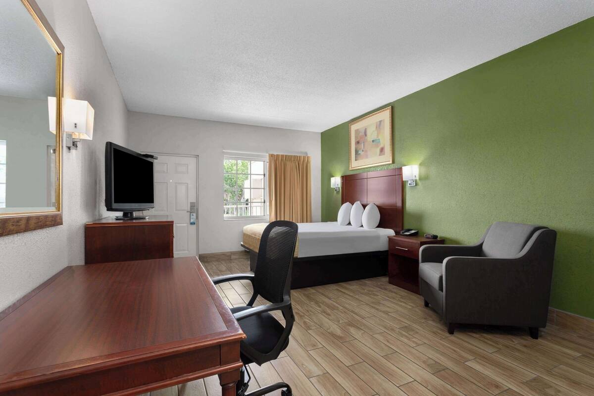 A hotel room with a wooden desk, a television, a bed with pillows, a chair, and a green wall.