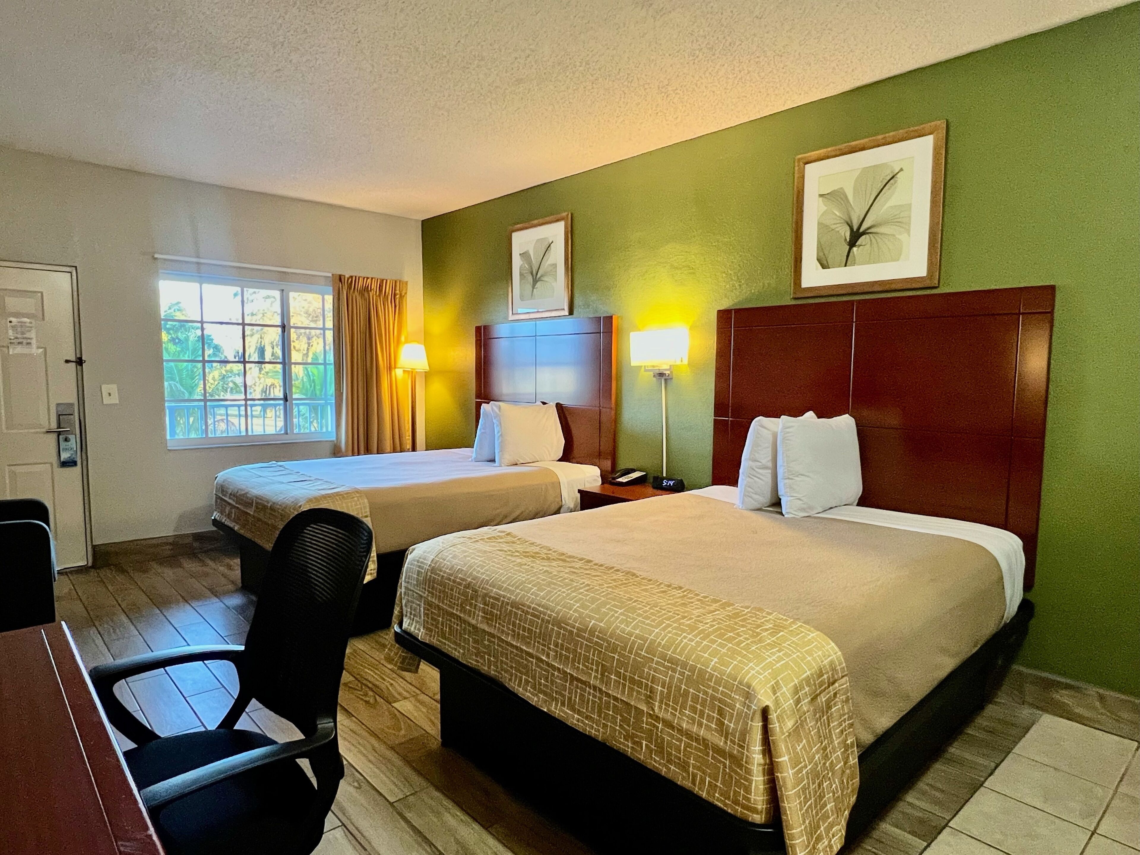A hotel room with two beds, a desk, and a chair. There are two bedside tables with lamps, a window with curtains, and a door.