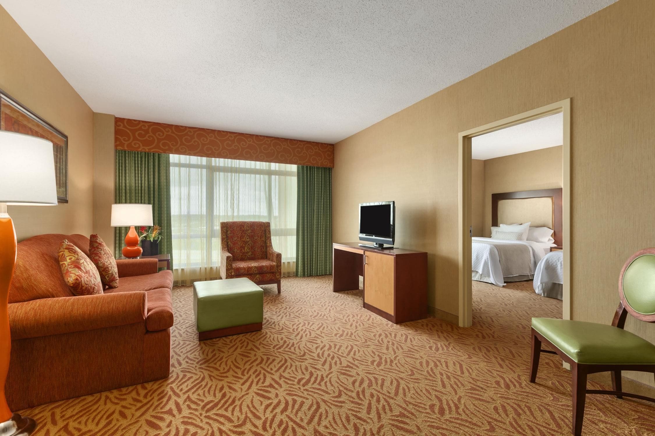 A hotel room with a sofa, armchair, ottoman, TV stand, and a bed.