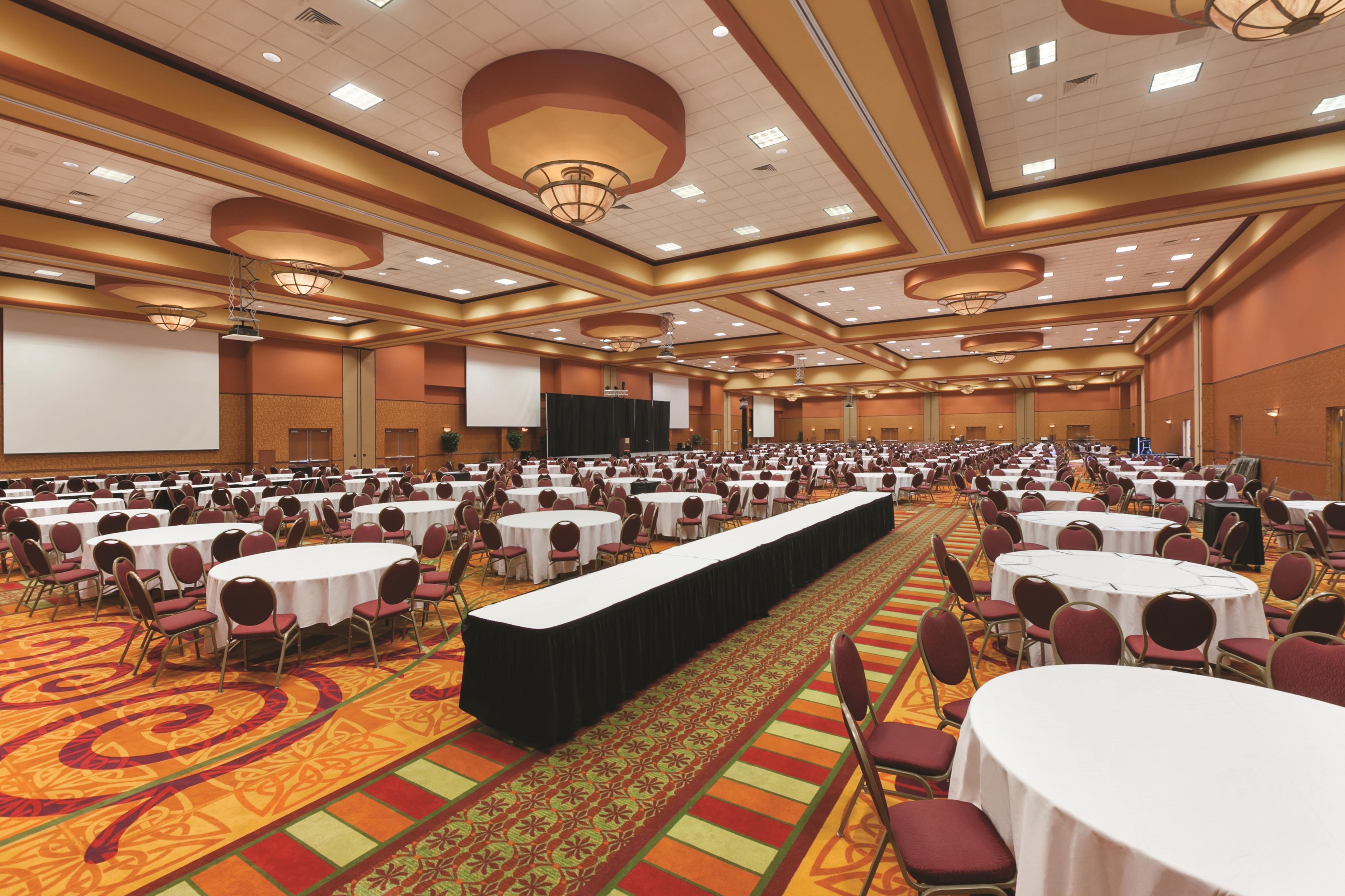 A large banquet hall with rows of round tables and chairs, a long table with a black tablecloth, and a stage with a screen.