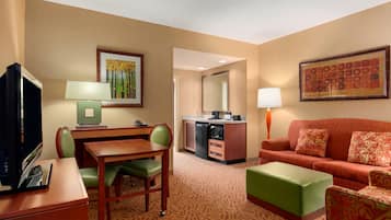 A hotel room with a dining table, green chairs, a sofa, a TV, a kitchen area, and a large framed picture on the wall.