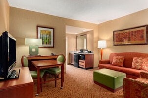 A hotel room with a dining table, green chairs, a sofa, a TV, a kitchen area, and a large framed picture on the wall.