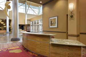 A hotel reception area with a curved wooden reception desk, a framed abstract painting on the wall, and a large open space with a high ceiling and natural light.