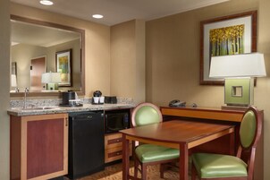 A hotel room with a kitchenette, a dining table with chairs, a mirror, a painting on the wall, and a lamp.
