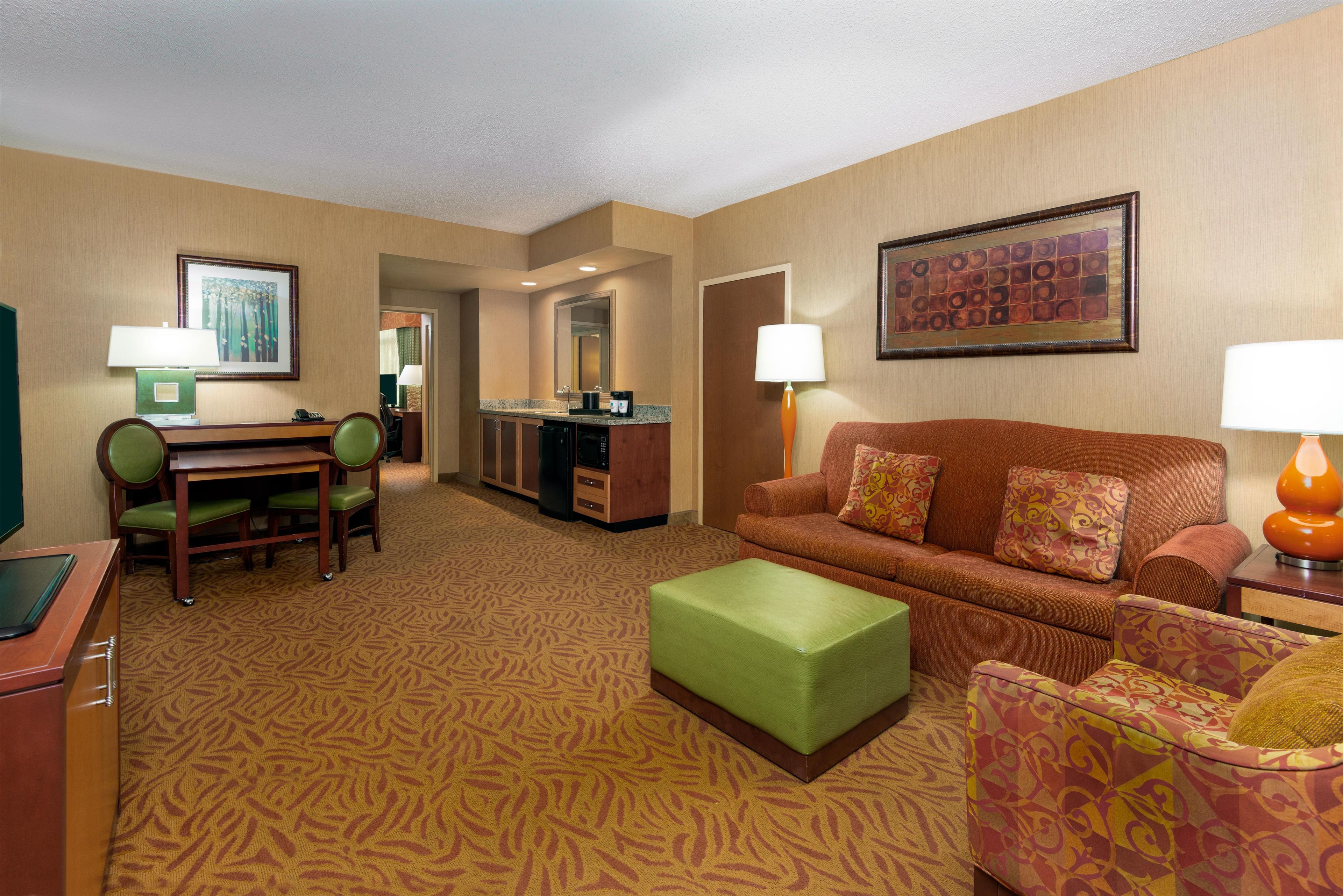 A hotel room with a green sofa, a green ottoman, a wooden desk with a chair, a kitchenette, and a large framed artwork on the wall.