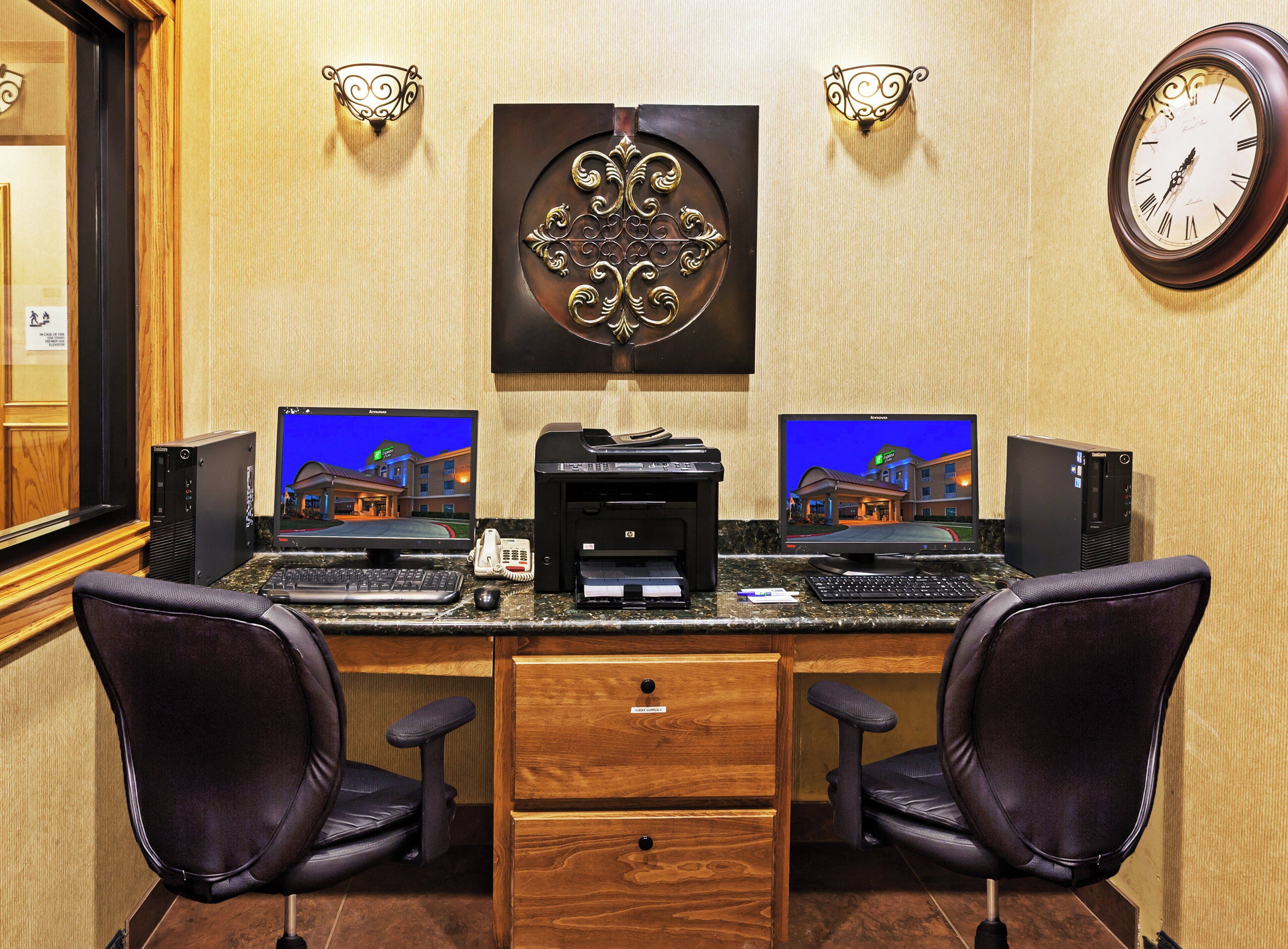 A hotel reception desk with two computer monitors, a telephone, a printer, and a clock on the wall.