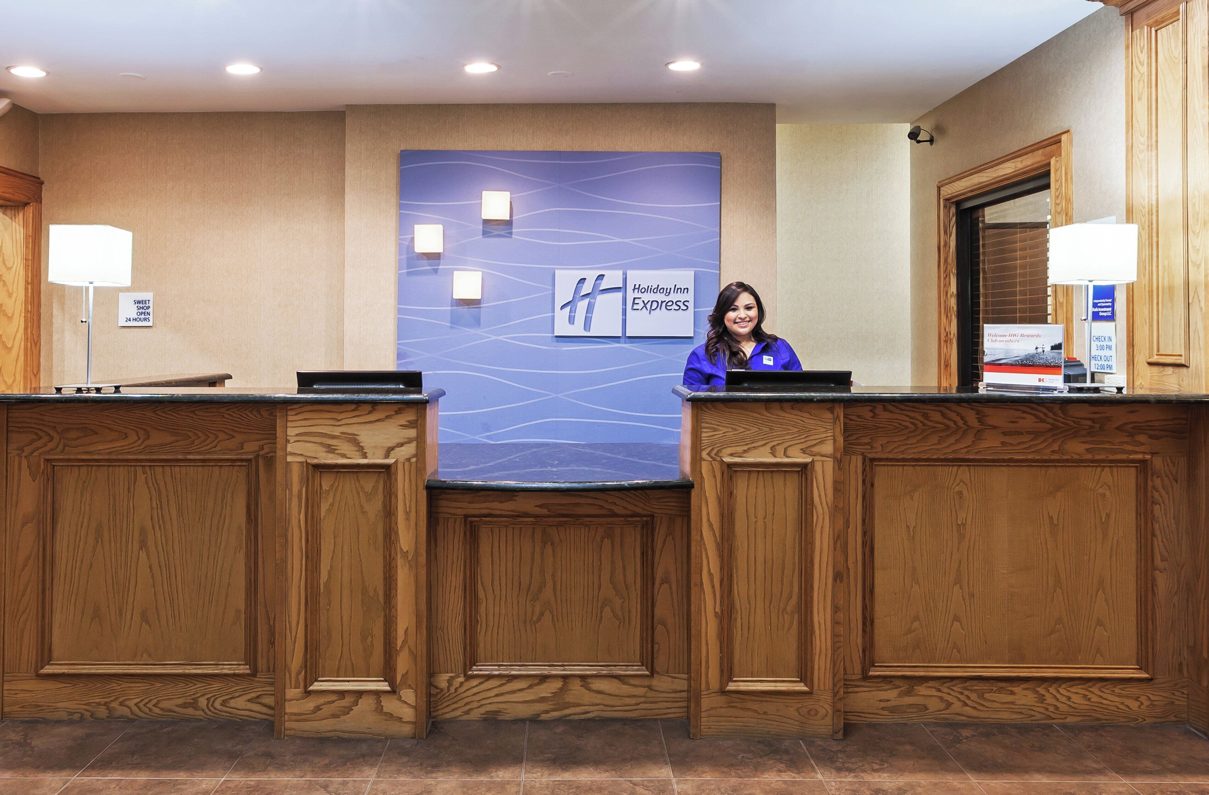 Un mostrador de recepción de hotel con una pared azul que muestra el logotipo de Holiday Inn Express, dos recepcionistas y un cartel que indica una tienda de dulces abierta las 24 horas.