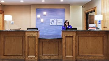 Un mostrador de recepción de hotel con una pared azul que muestra el logotipo de Holiday Inn Express, dos recepcionistas y un cartel que indica una tienda de dulces abierta las 24 horas.