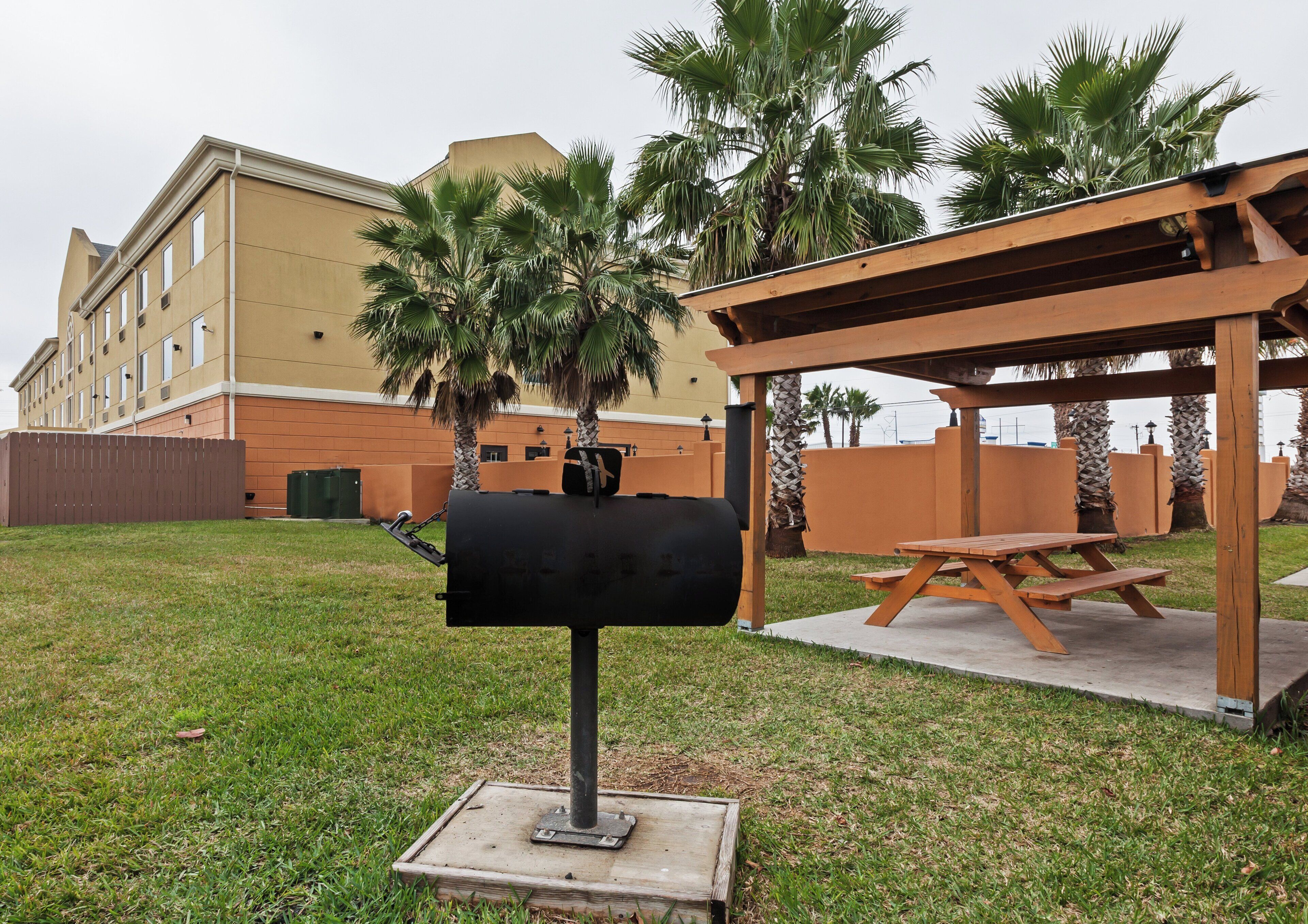 Una parrilla con una mesa y bancos de madera, rodeada de césped y palmeras, con un edificio al fondo.