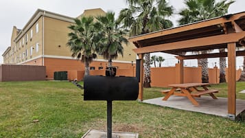 Una parrilla con una mesa y bancos de madera, rodeada de césped y palmeras, con un edificio al fondo.