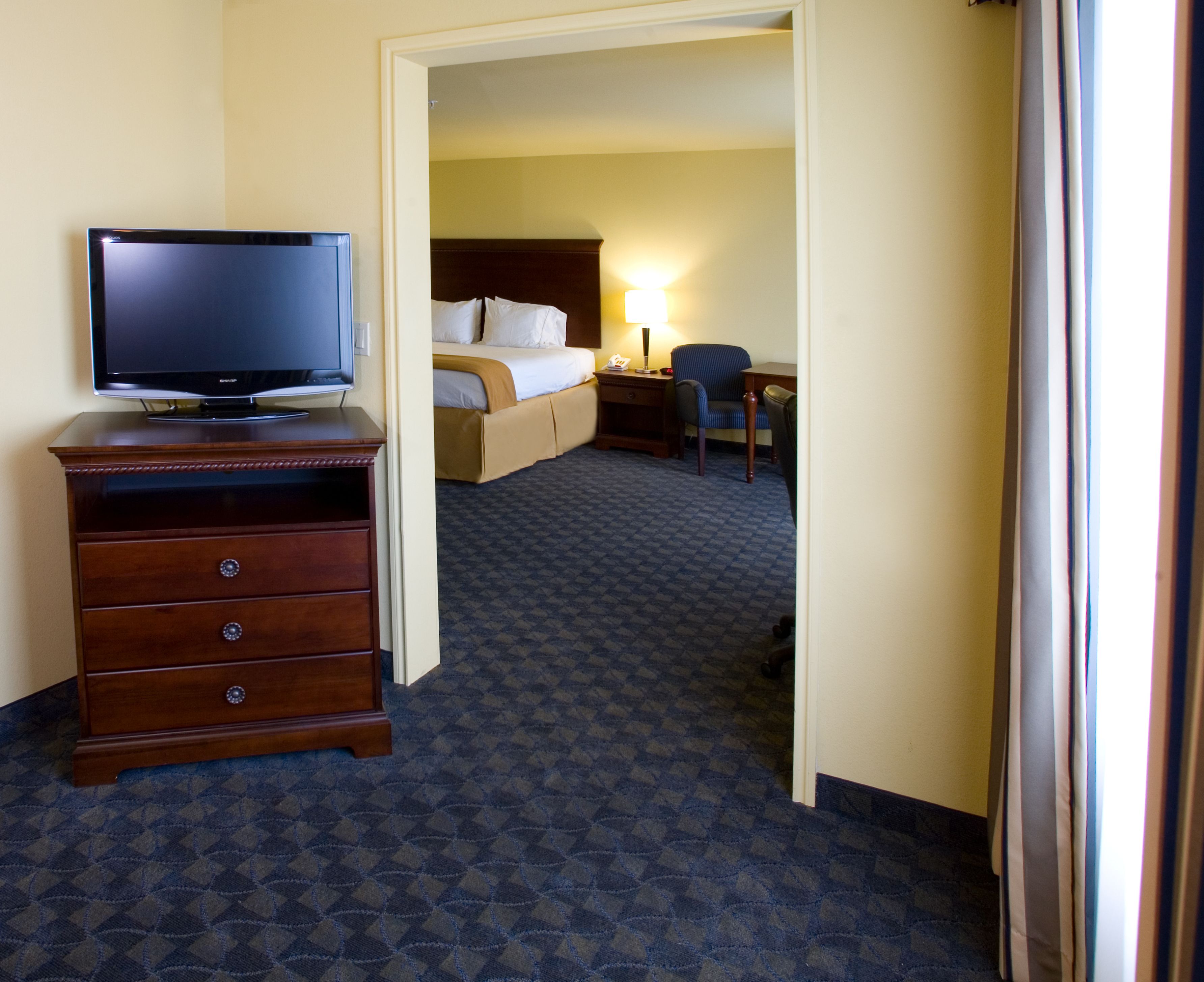 A hotel room with a flat-screen TV on a wooden dresser, a bed with white linens, a desk with a chair, and a patterned carpet.